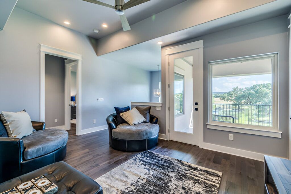 A modern living room featuring a round, cushioned chair, a sleek leather armchair, and a patterned rug. Large windows open to a scenic balcony view of lush greenery. The room has stylish decor with recessed lighting, a ceiling fan, and warm hardwood flooring, showcasing contemporary interior design ideas.