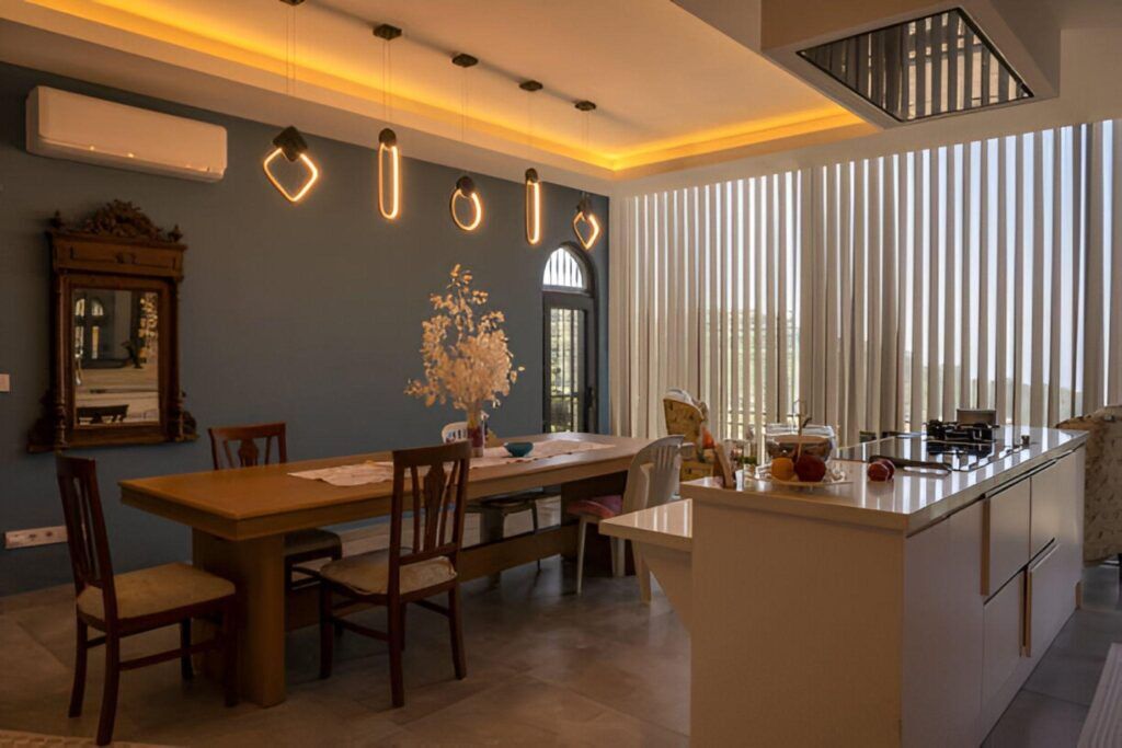 Modern kitchen interior with unique pendant lights and warm ambient lighting, featuring a wooden dining table, classic wooden chairs, and tall blinds covering large windows.