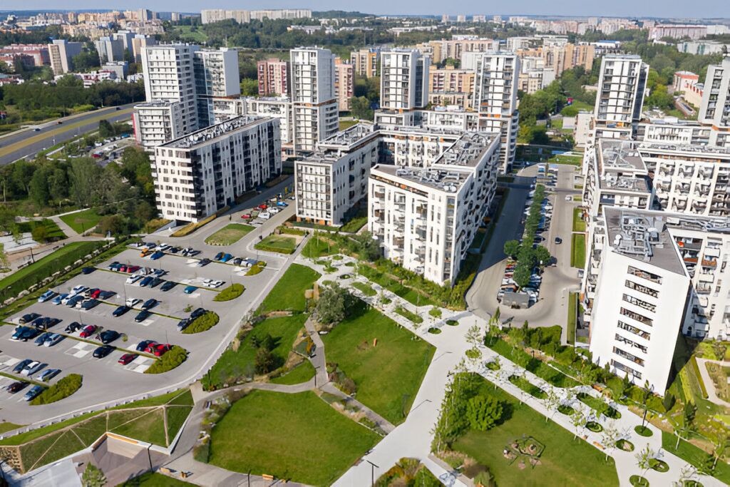 A modern urban residential complex featuring multiple tall buildings surrounded by green spaces, walkways, and a parking lot, located in a bustling cityscape.